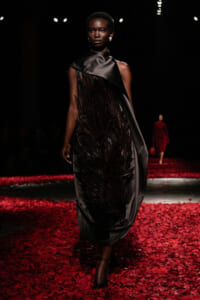 Model walking a runway in a black satin dress with feathered centerpiece, surrounded by red petals on the floor.