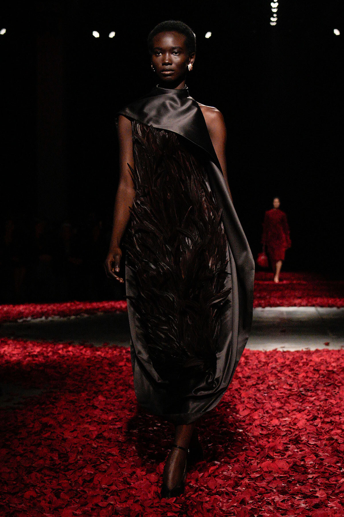Model walking a runway in a black satin dress with feathered centerpiece, surrounded by red petals on the floor.