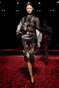 Model walking a runway in a sheer black-and-white printed dress with belt, carrying a gray clutch on a red petal-covered floor