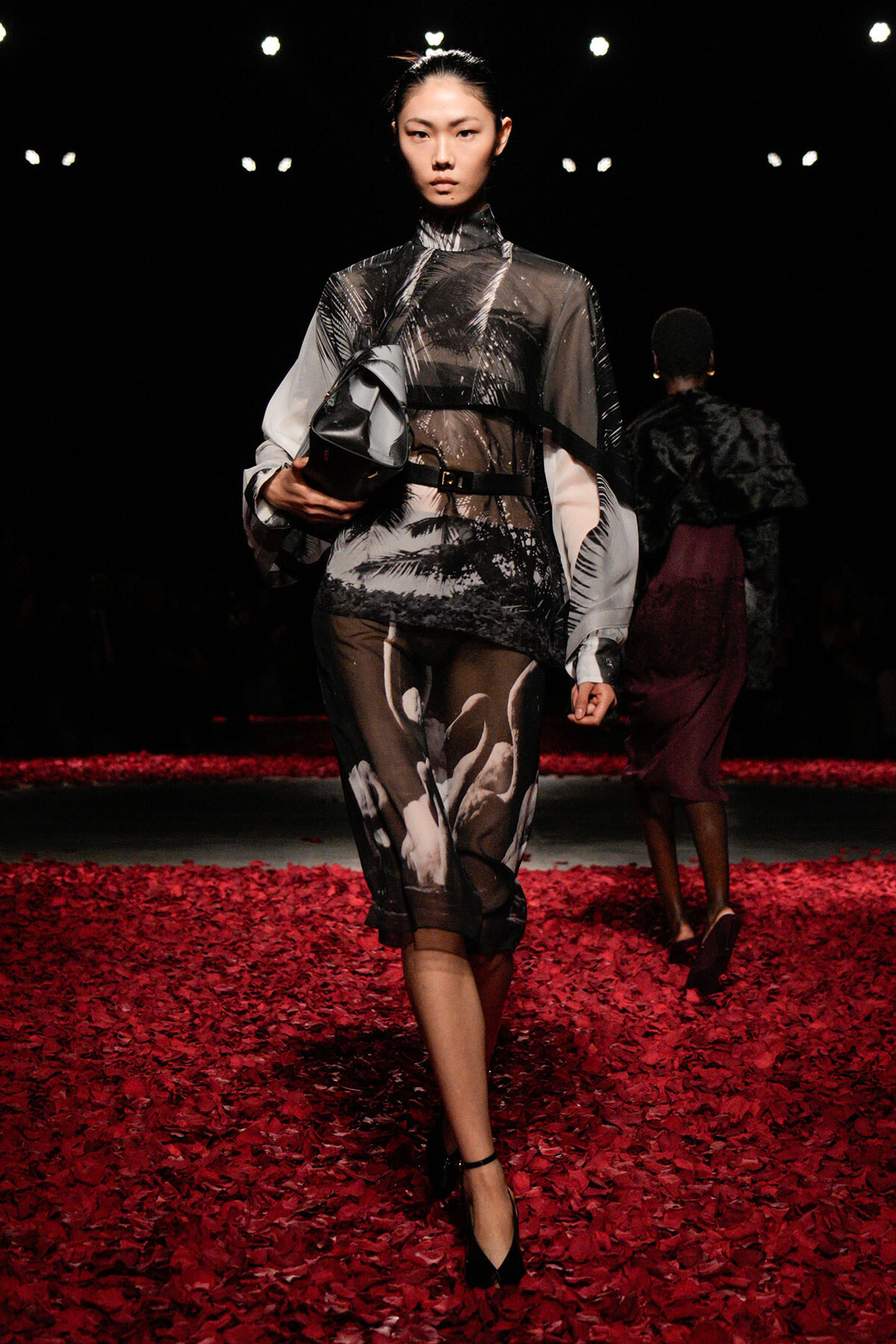 Model walking a runway in a sheer black-and-white printed dress with belt, carrying a gray clutch on a red petal-covered floor