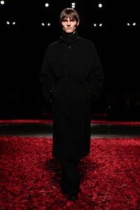Model walking a runway in a long black belted coat, hands in pockets, on a stage scattered with red petals.