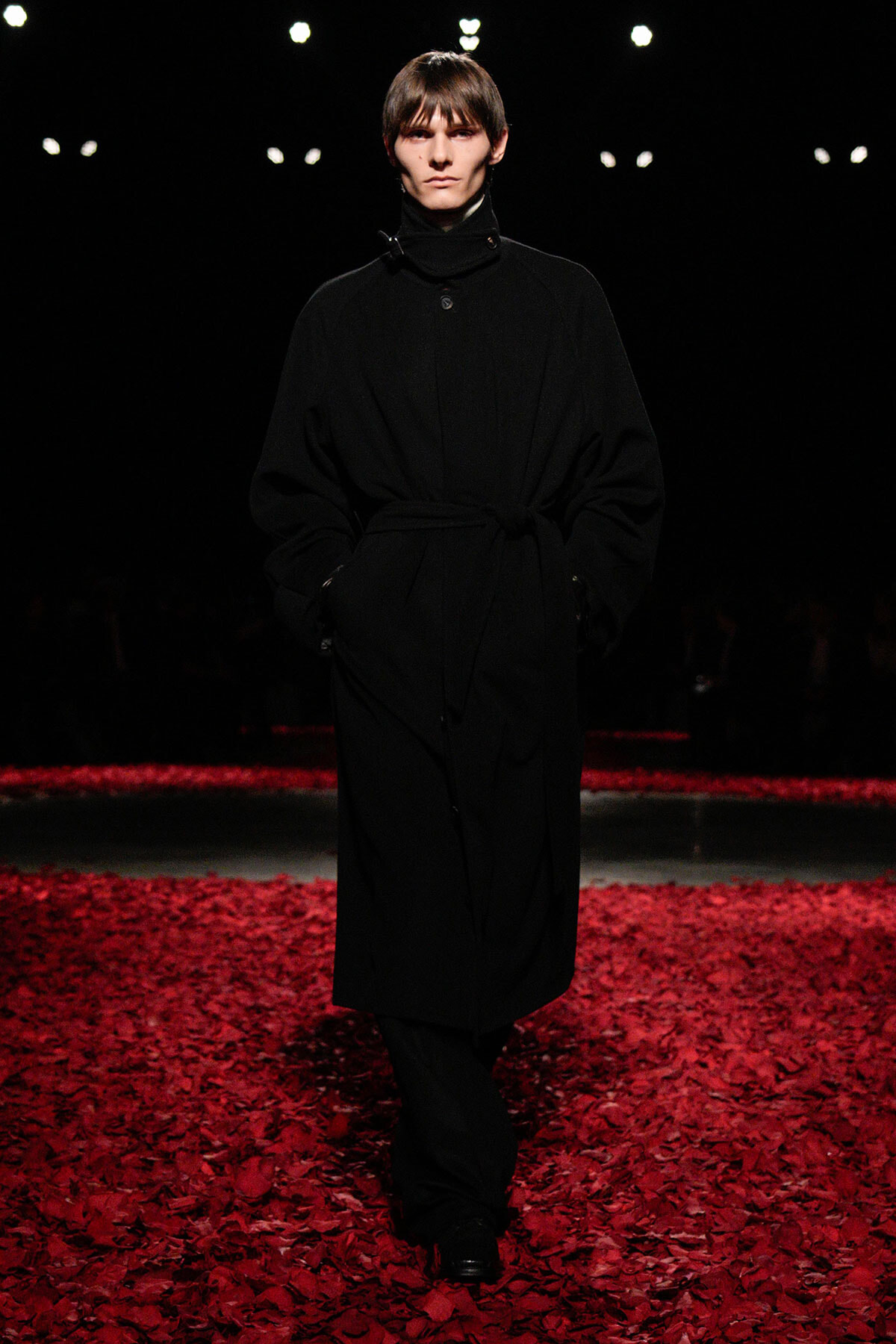 Model walking a runway in a long black belted coat, hands in pockets, on a stage scattered with red petals.