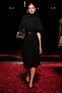 Model walking the runway in a black cape-dress, belt, and pointed heels, holding a black clutch. Red petals cover the floor.