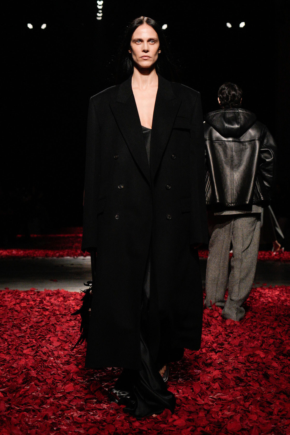 Model on a runway wearing a long black double-breasted coat over a black outfit; red petals cover the floor. A second model in a leather jacket walks away in the background.