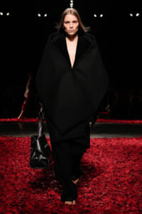 Model walks the runway in an oversized black cape coat, barefoot, carrying a black handbag on a red petal-covered floor.