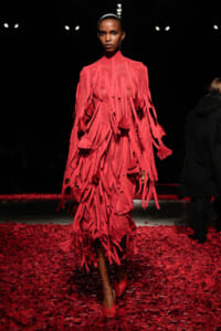Model walks the runway in a vivid pink fringe dress with tassels, standing against a dark backdrop; floor covered in red petals.