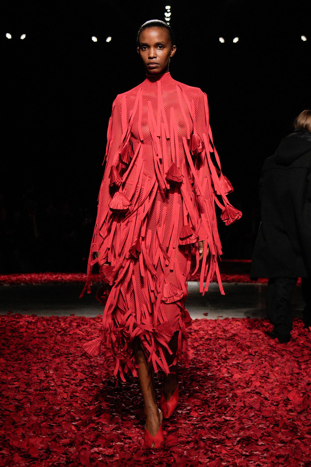 Model walks the runway in a vivid pink fringe dress with tassels, standing against a dark backdrop; floor covered in red petals.