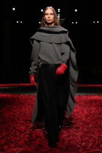 Model on a runway wearing a gray layered cape over black pants with red gloves, walking over a floor scattered with red petals.