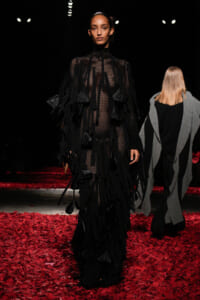 Model walking a runway in a long black fringed mesh dress, surrounded by red petals on the floor under dark lighting.