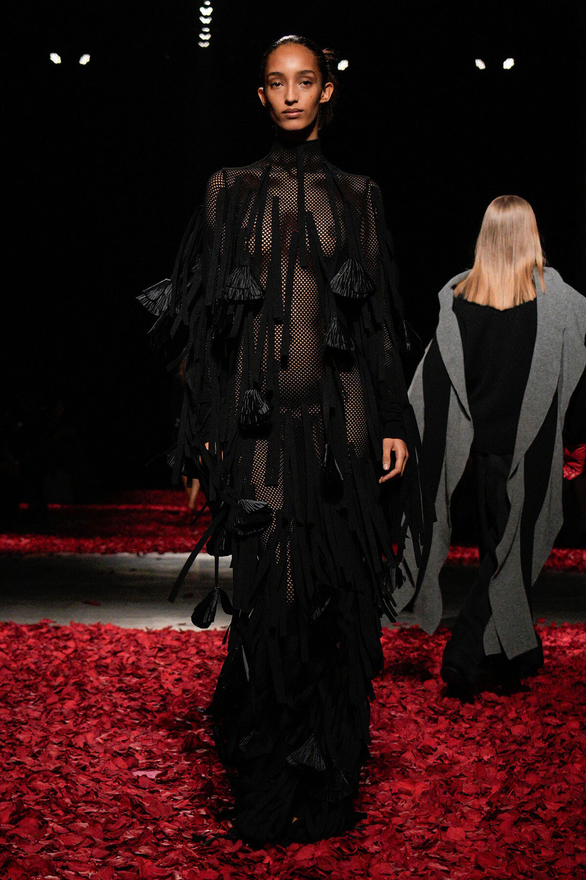 Model walking a runway in a long black fringed mesh dress, surrounded by red petals on the floor under dark lighting.