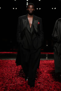 Male model walking a fashion runway in an oversized black suit with a deep V-neck and red pocket square, surrounded by red rose petals on the floor.