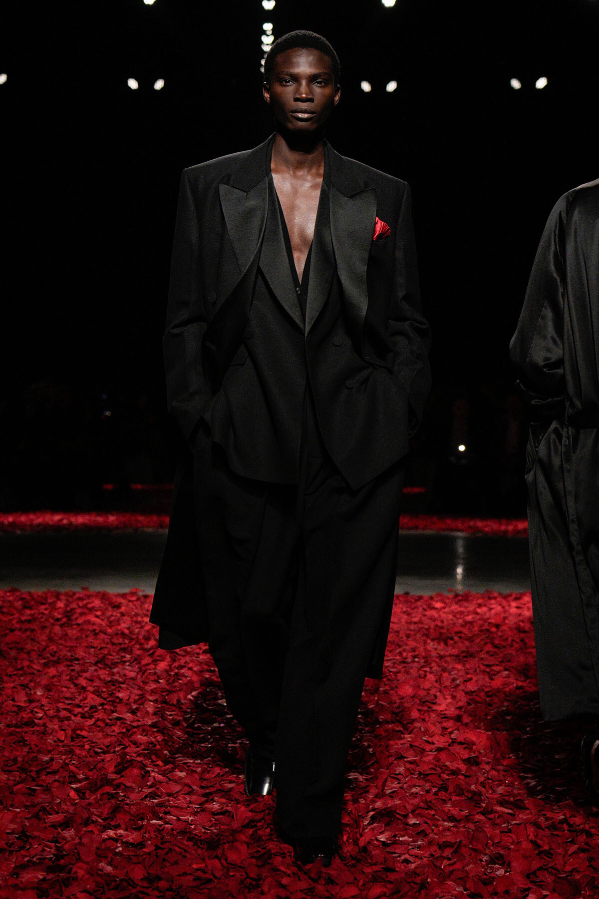 Male model walking a fashion runway in an oversized black suit with a deep V-neck and red pocket square, surrounded by red rose petals on the floor.