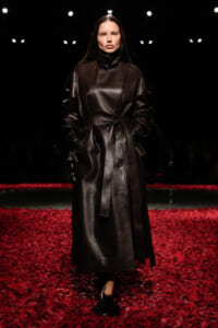 Model walking a runway in a long black leather trench coat, belted at the waist, on a floor covered with red petals.