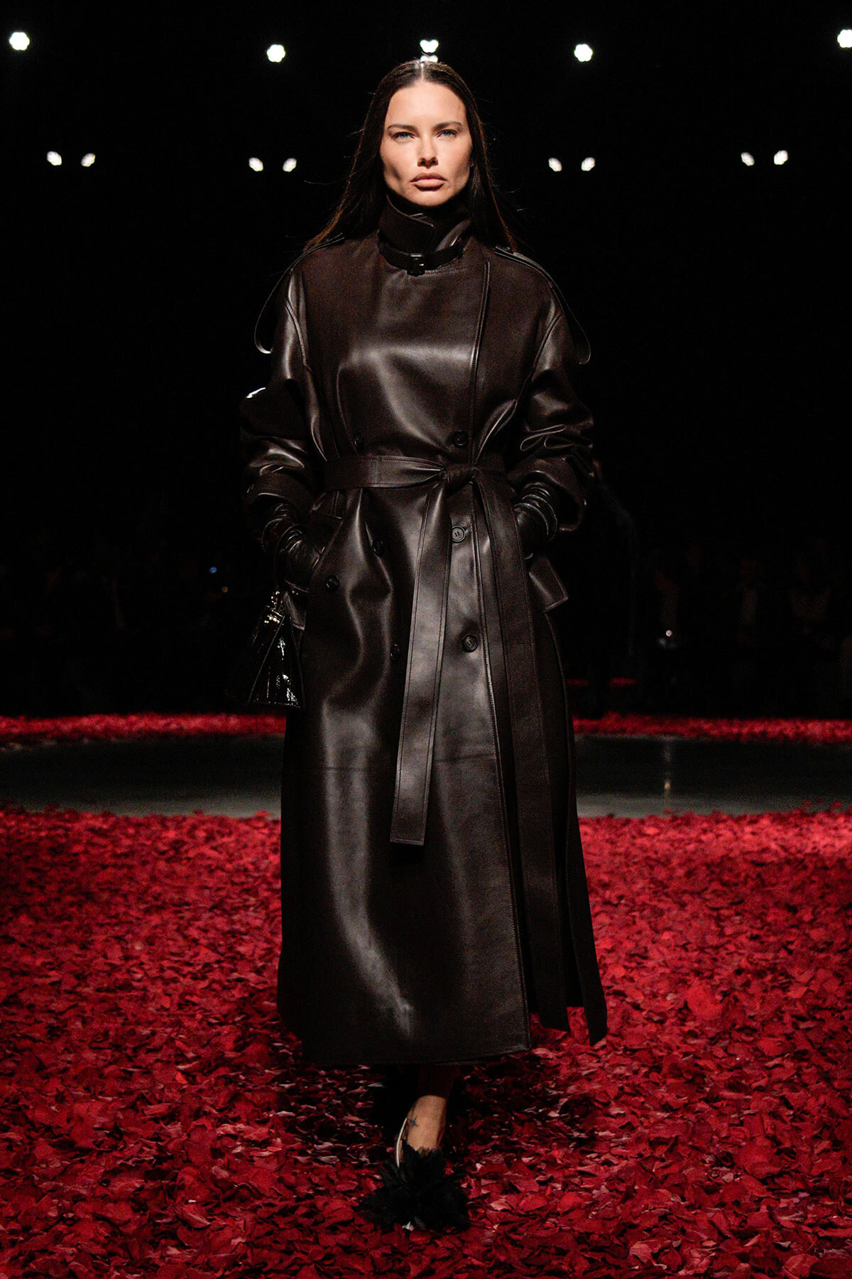 Model walking a runway in a long black leather trench coat, belted at the waist, on a floor covered with red petals.