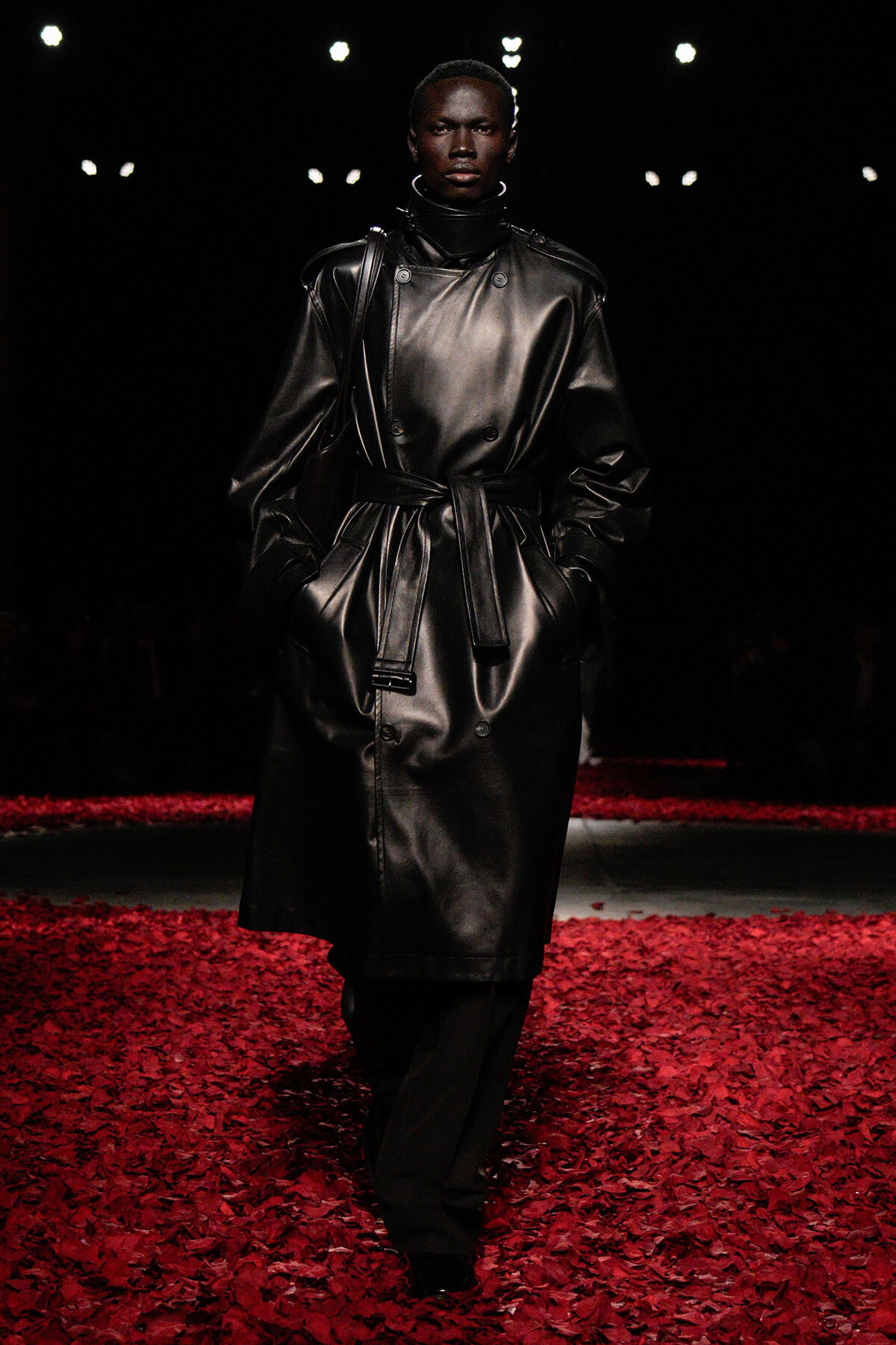 Model walking a runway in a black leather trench with belt, hands in pockets, over a red petal floor under spotlights