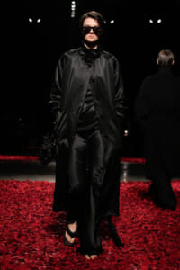 Model in a long black satin coat and oversized sunglasses walks a dim runway, red petals on the floor beneath her.