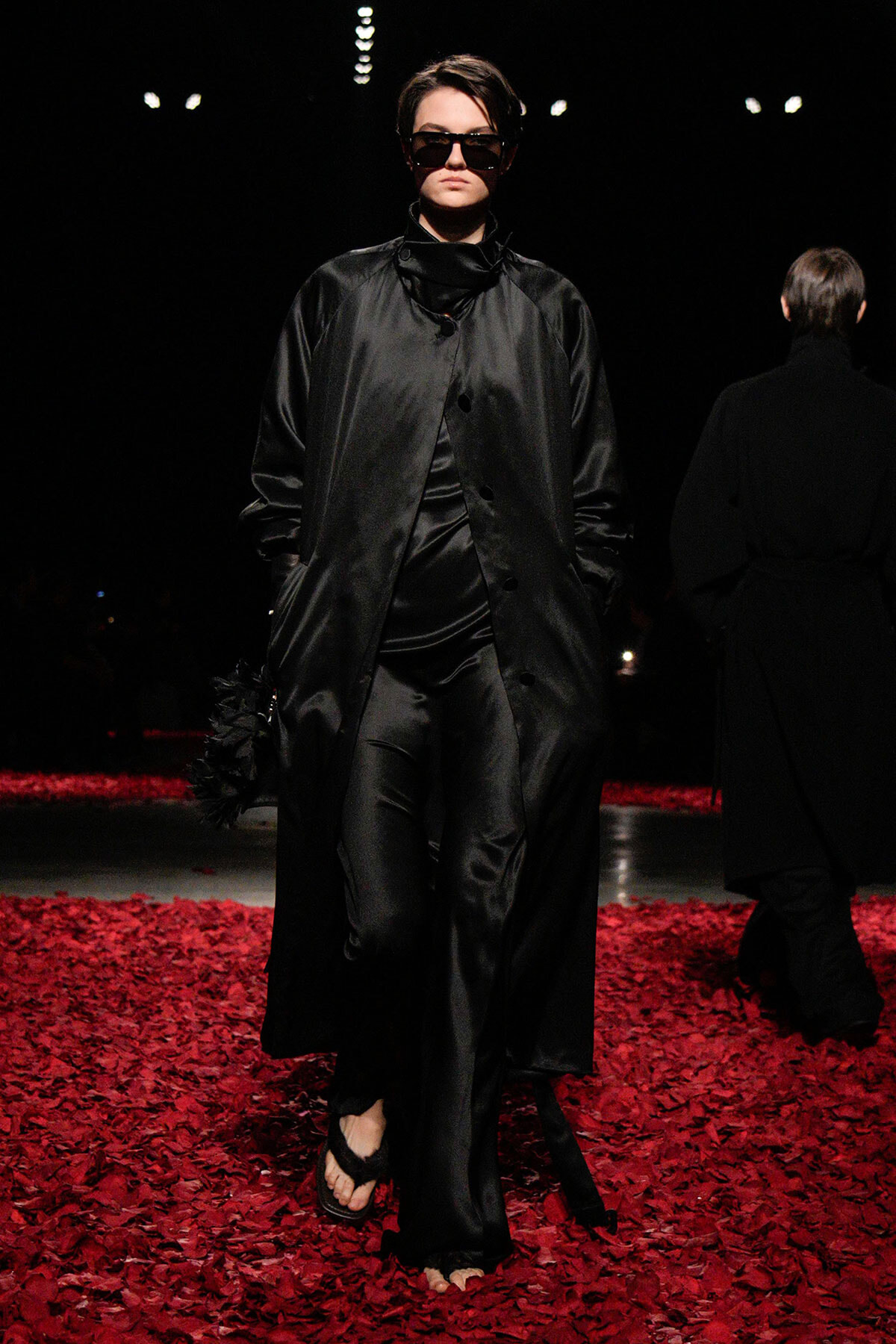 Model in a long black satin coat and oversized sunglasses walks a dim runway, red petals on the floor beneath her.