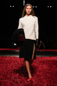 Model walking a fashion show on a red rose-petal runway, wearing a white sweater and black slit-skirt, carrying a black coat and burgundy handbag with gloves.