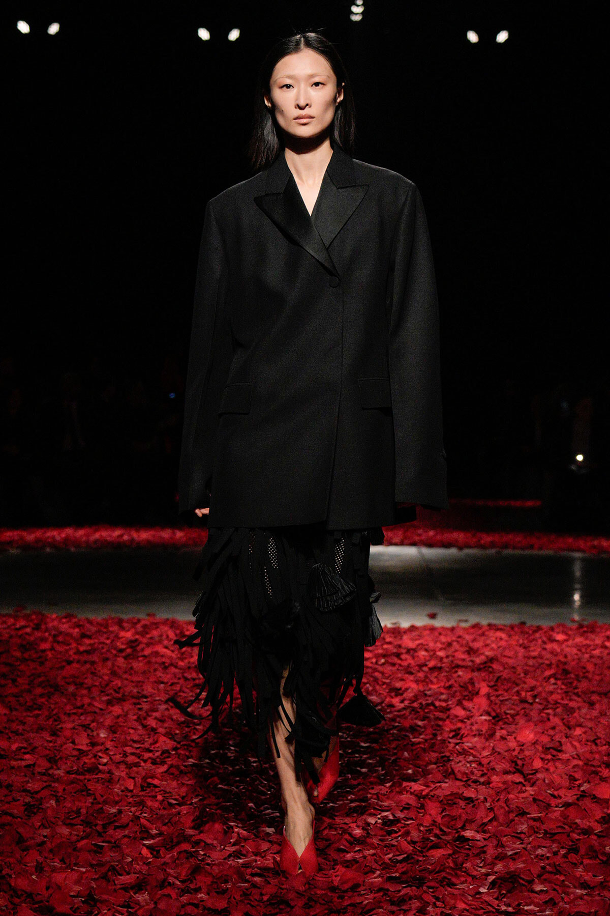 Model walking a runway in an oversized black blazer with a fringed black skirt and red heels, surrounded by red petals on the floor.