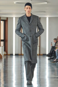 Male model walks a runway wearing a heather gray wool coat, black sweater, dark pants, and a black beanie.
