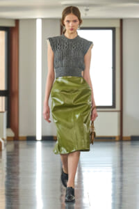 Model walks a runway in a gray cable-knit sleeveless top and high-waisted olive leather skirt, black shoes, and a small handbag.