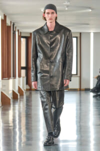 Male model on a fashion runway wearing a glossy black leather coat and matching leather pants, with a dark knit cap.