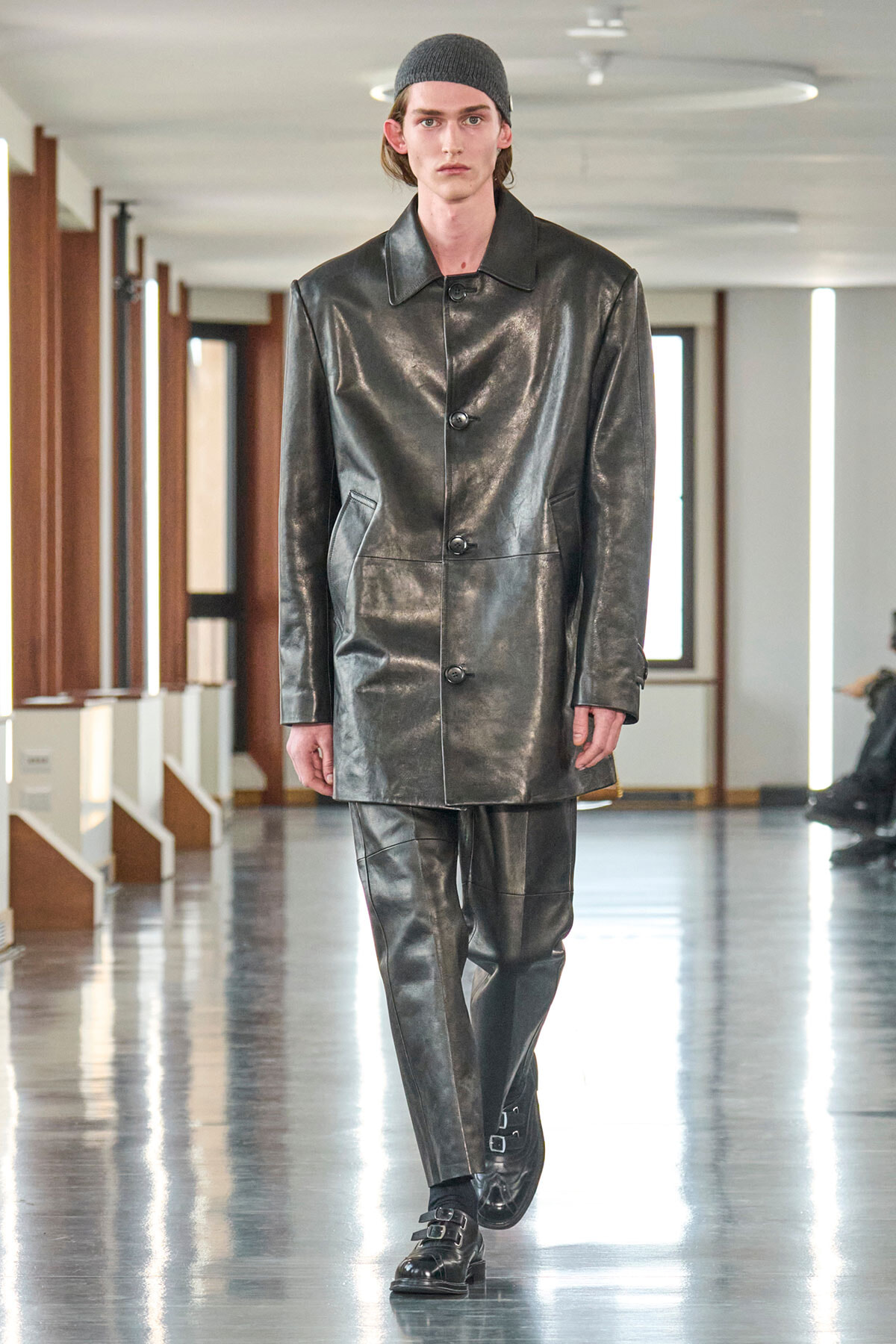 Male model on a fashion runway wearing a glossy black leather coat and matching leather pants, with a dark knit cap.