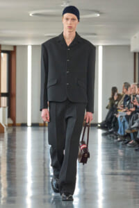 Male model walking a runway in a dark tailored suit with a buttoned jacket, black loafers, and a navy beanie, carrying a small brown strap bag.