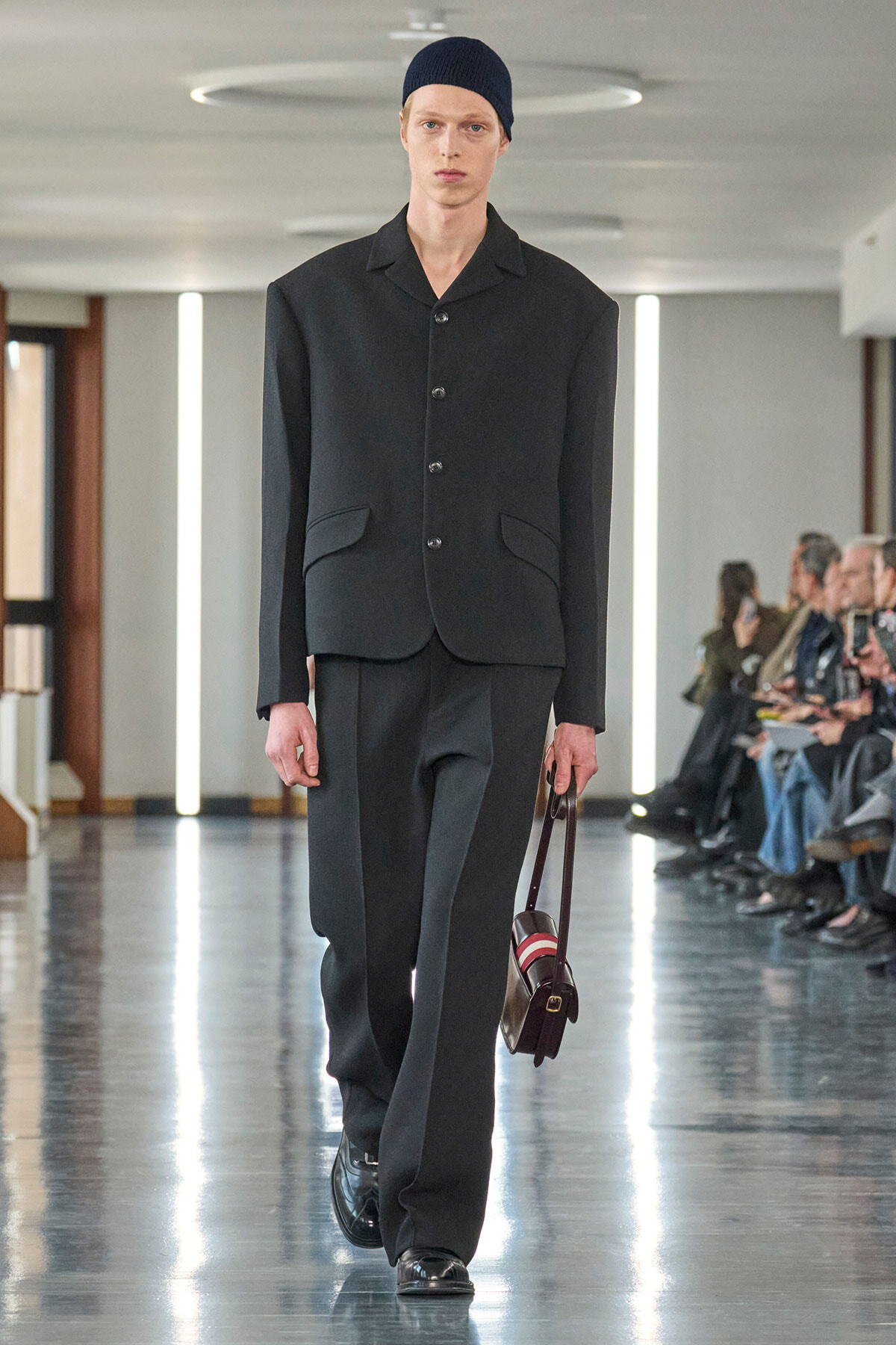 Male model walking a runway in a dark tailored suit with a buttoned jacket, black loafers, and a navy beanie, carrying a small brown strap bag.
