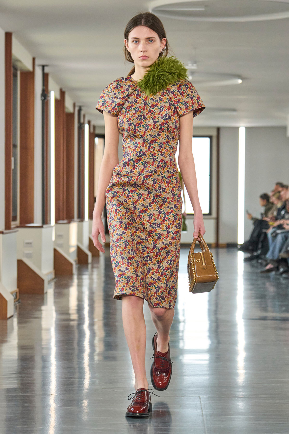Model walking a runway in a colorful floral midi dress with a green feather accent at the shoulder, carrying a small brown handbag.