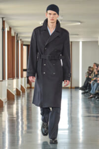 Male model walks a runway in a double-breasted black wool trench with a belt and black beanie.