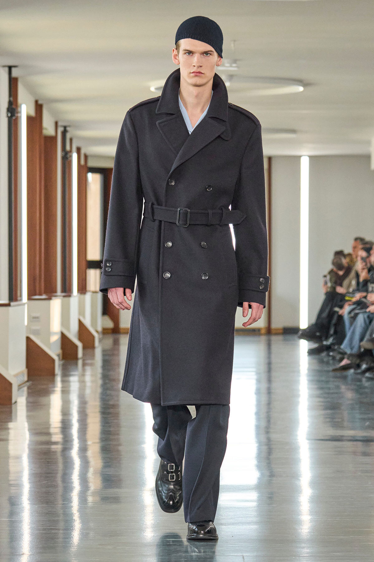 Male model walks a runway in a double-breasted black wool trench with a belt and black beanie.