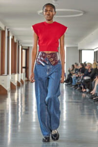 Model walking a runway in a red cropped knit top, oversized blue jeans, and a wide burgundy belt with multiple buckles cinching the waist; audience visible along the catwalk in the background.