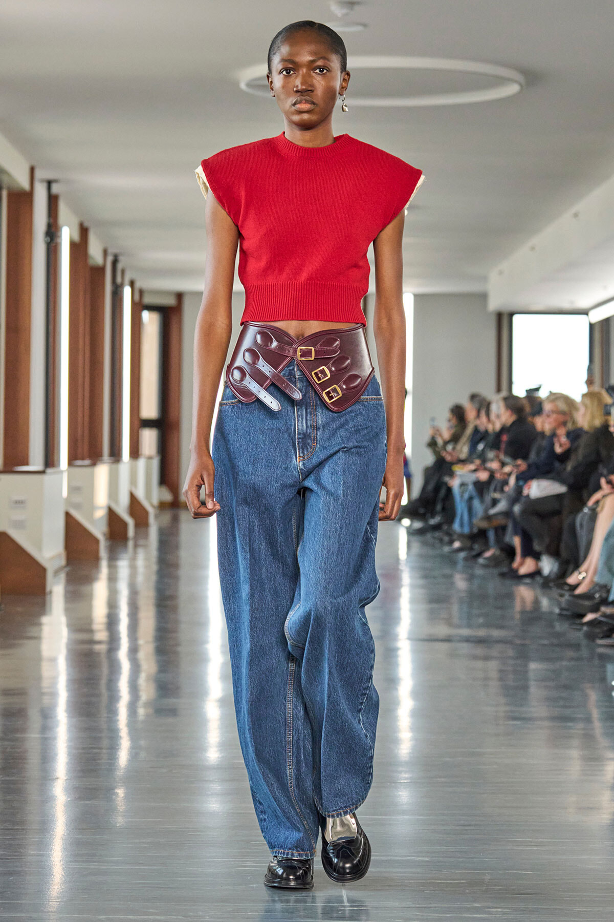 Model walking a runway in a red cropped knit top, oversized blue jeans, and a wide burgundy belt with multiple buckles cinching the waist; audience visible along the catwalk in the background.