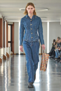 Female model walking a denim-on-denim outfit down a runway, carrying a floral tote bag.