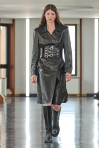 Model walking the runway in a black leather trench dress with a wide three-buckle belt and knee-high boots.