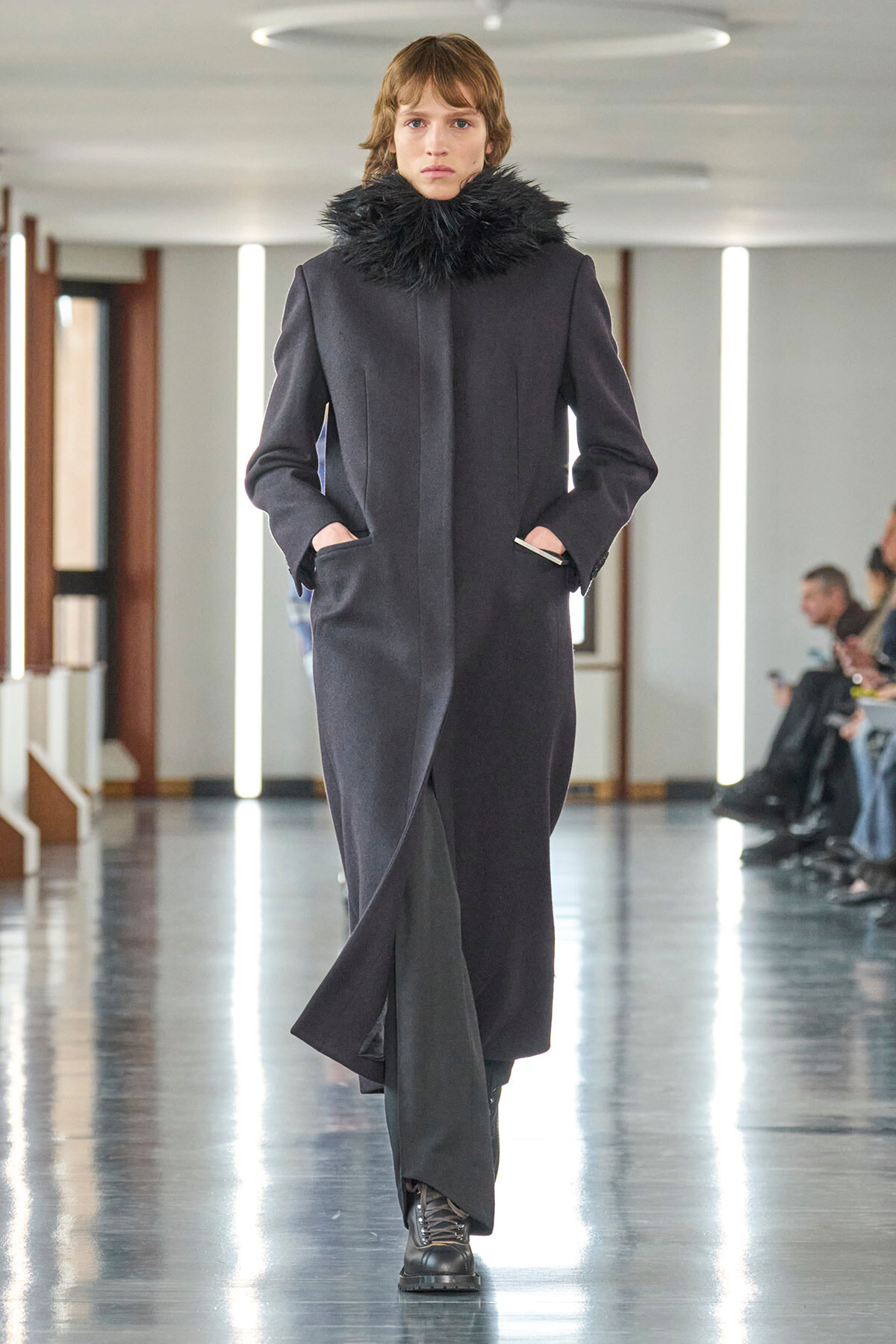 Female model walking a runway in a dark long coat with a black fur collar, hands in pockets.