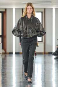 Model walking a runway in an oversized black leather jacket and tailored black pants on a glossy floor runway.