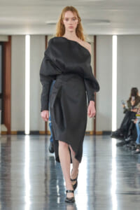 Model walks a runway in a black one-shoulder midi dress with a voluminous sleeve and asymmetric hem, black Mary Jane shoes.