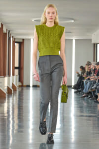Female model walks a fashion show in an olive-green sleeveless knit top and gray tailored pants, carrying a small green handbag.