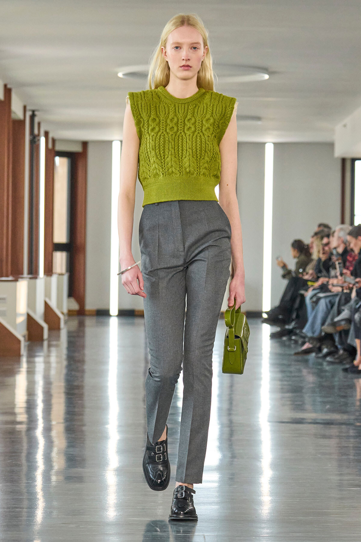 Female model walks a fashion show in an olive-green sleeveless knit top and gray tailored pants, carrying a small green handbag.