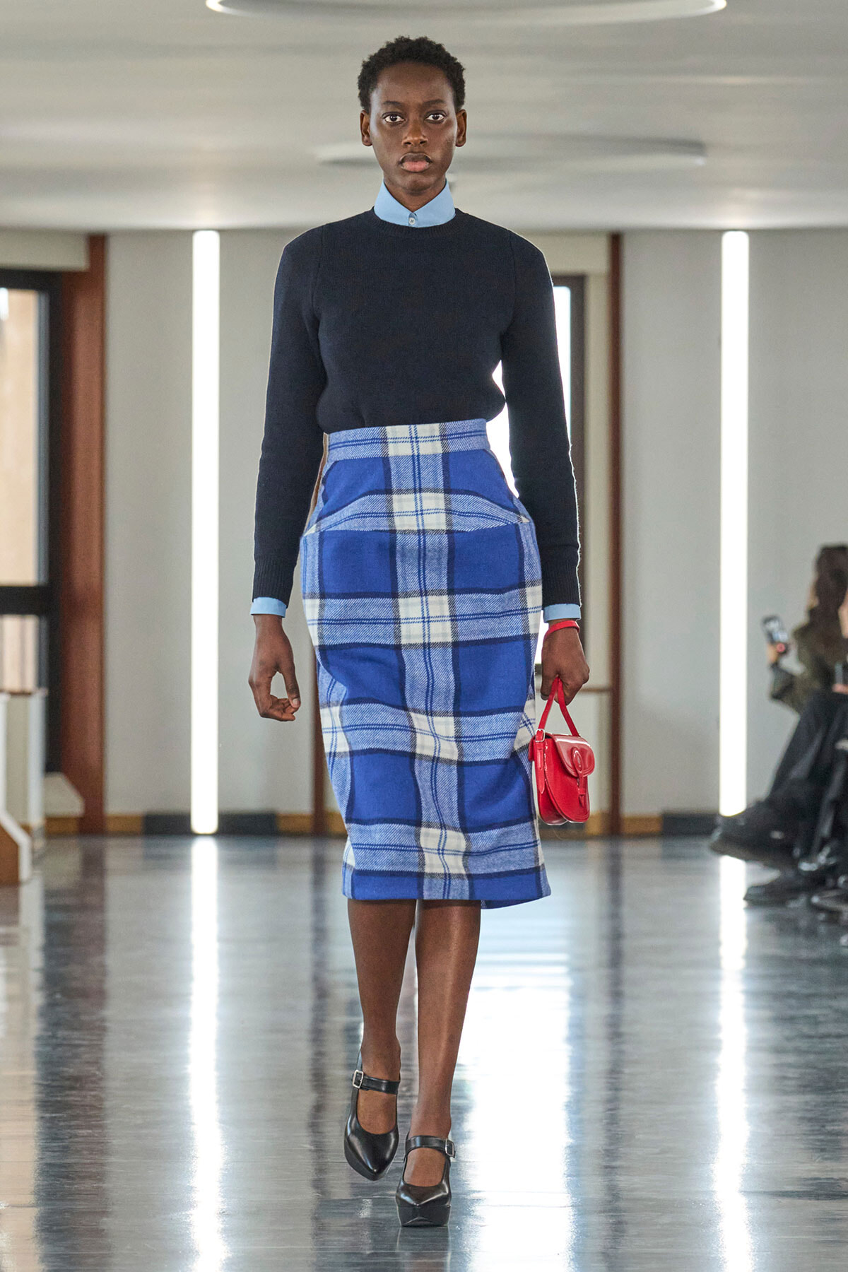 Model walking down a runway in a navy knit top with a light blue collar, a blue plaid midi skirt, black Mary Jane heels, and a small red handbag.