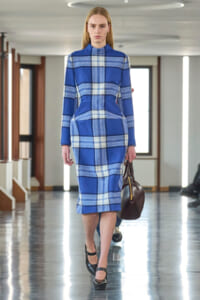 Model walking down a runway in a blue plaid midi dress with structured pockets, black heels, and a brown handbag.