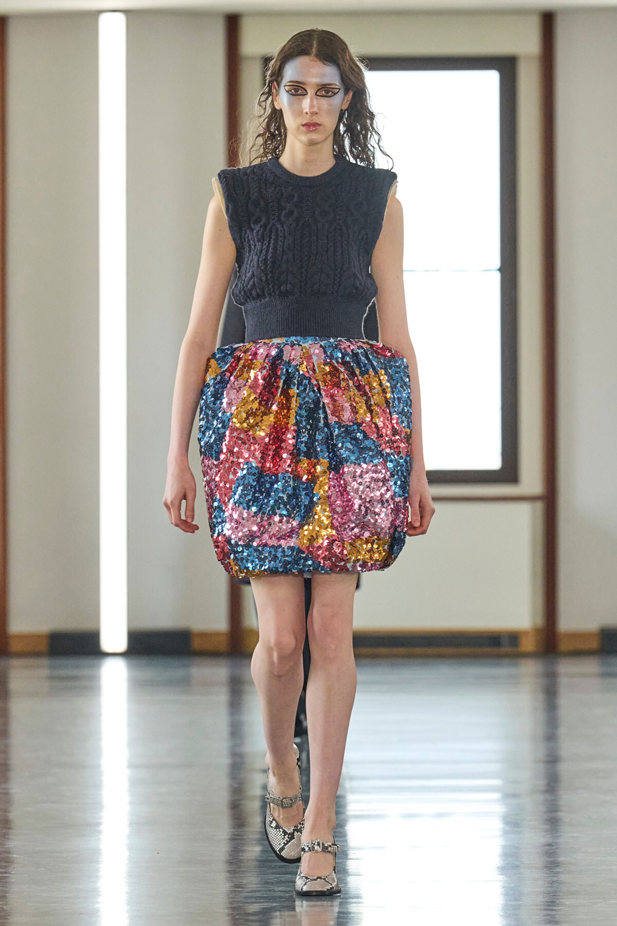 Model walking on a runway in a navy sleeveless knit top and a bright multicolored sequin bubble skirt with snakeskin flats.
