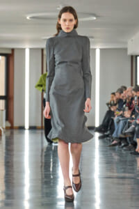 Model walking a runway in a gray high-neck long-sleeve dress with padded shoulders and a flared hem.