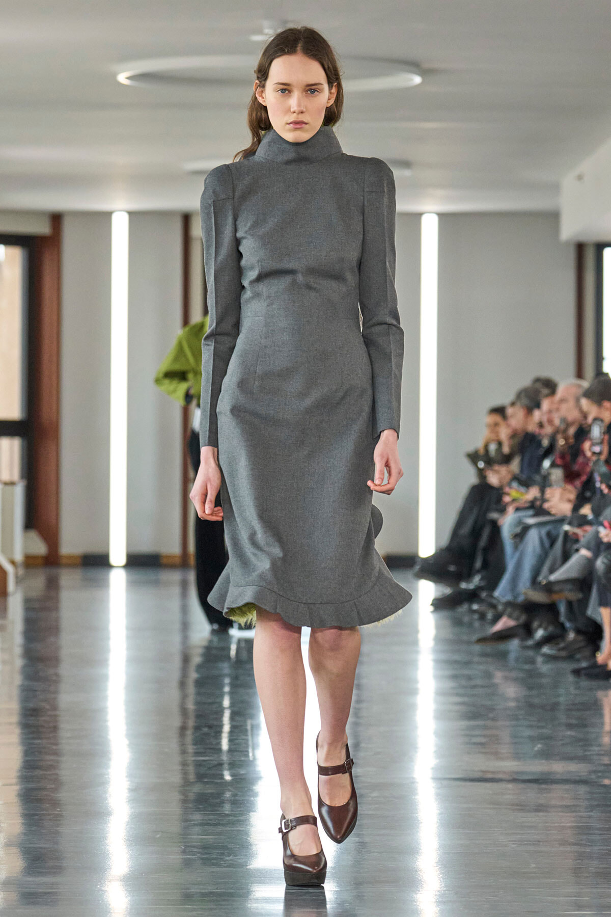 Model walking a runway in a gray high-neck long-sleeve dress with padded shoulders and a flared hem.