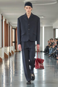 Male runway model in a navy suit and black beanie walks the catwalk, holding a red tote bag beside him.