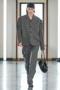 Model walks a runway in a loose gray suit with a matching blazer and trousers, black cap, and a black leather handbag.