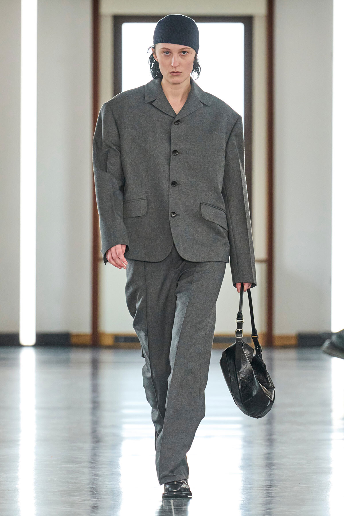 Model walks a runway in a loose gray suit with a matching blazer and trousers, black cap, and a black leather handbag.
