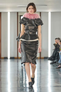 Female model walking runway in a black leather peplum top and matching pencil skirt with a pink feathered collar, black lace-up shoes.